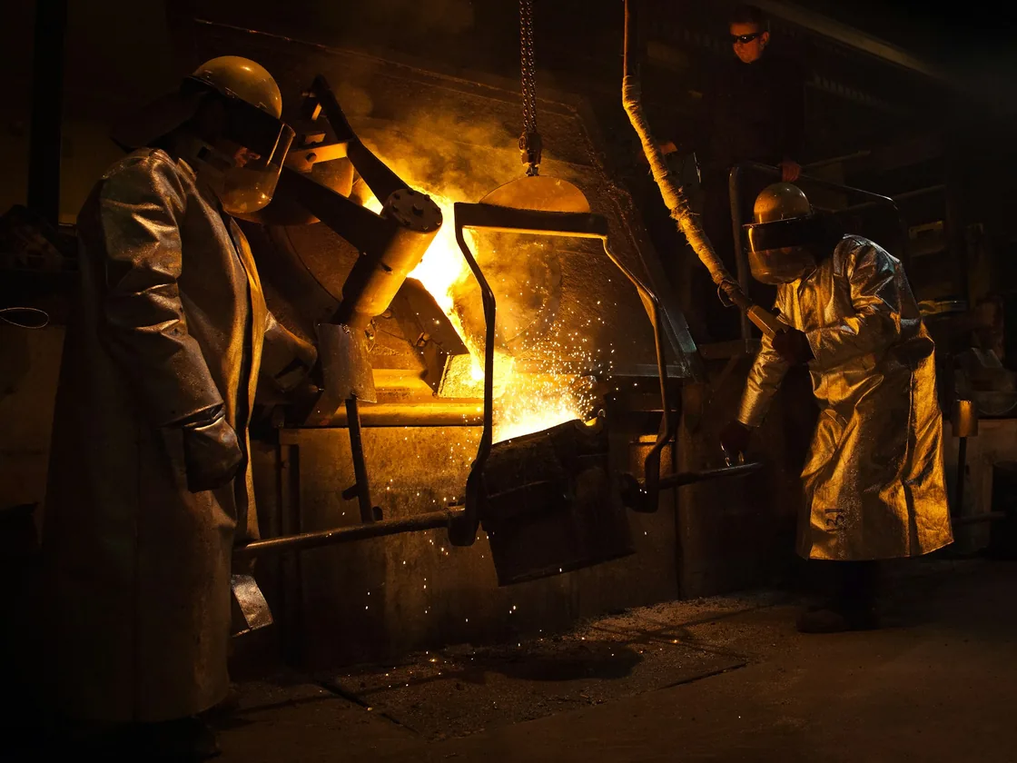Dos personas vierten metal fundido desde un crisol en un taller de fundición